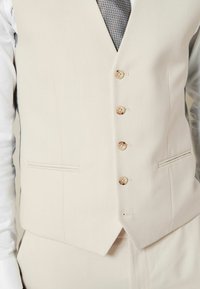 Un gilet de couleur crème en tissu texturé. Il présente cinq boutons en bois et deux poches poitrine, associé à une cravate grise à motifs.