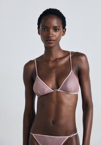 Lichtroze lingerieset met een driehoekige bralette, dunne bandjes en een decoratieve zilverkleurige hardware-accent in het midden.