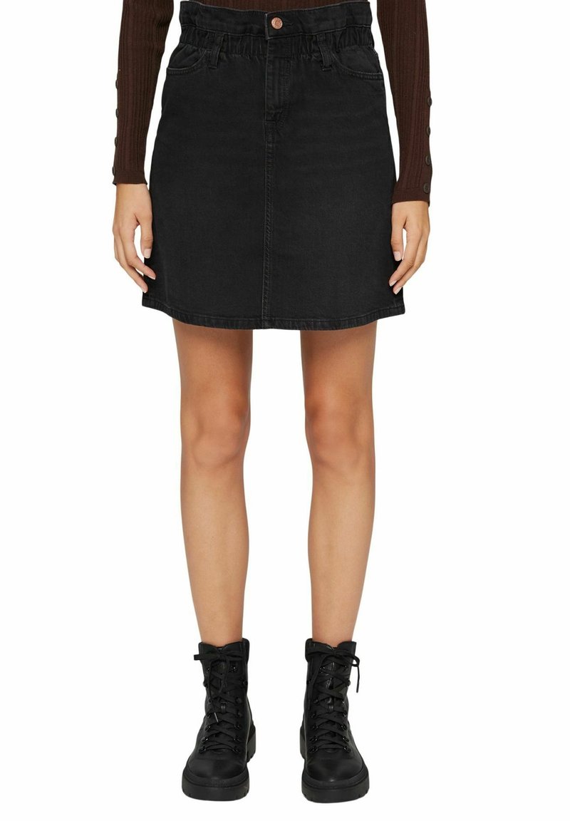 Femme portant une mini-jupe en denim noir avec des poches avant et un top marron côtelé à manches longues, associée à des bottines noires à lacets.