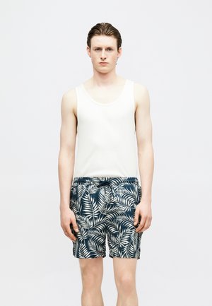 Νέος άνδρας όρθιος, φοράει λευκό αμάνικο πουκάμισο και σκούρο μπλε σορτς με λευκό τροπικό φυλλώδες μοτίβο.