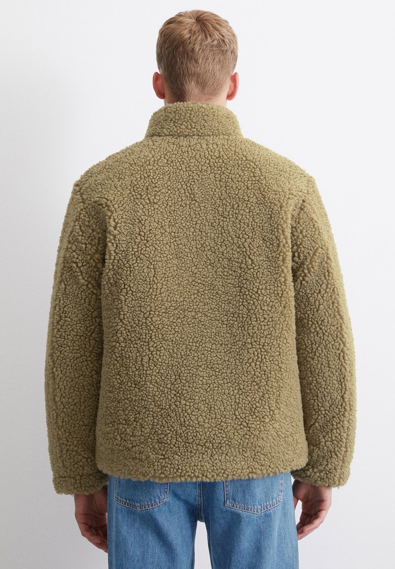 Sherpa-Jacke in Olivgrün mit hohem Kragen und strukturiertem Stoff. Lange Ärmel mit einer legeren Passform, kombiniert mit hellblauen Jeans.