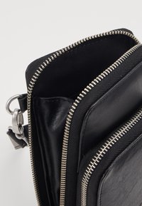 Musta vetoketjullinen laukku, joka on valmistettu mokasta ja kankaasta. Laukussa on hopeiset yksityiskohdat ja teksturoitu pinta. Sen avoin yläosa paljastaa sisävuorauksen.