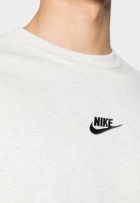 Jasnoszara bawełniana koszulka z czarnym haftowanym logo Nike po lewej stronie klatki piersiowej. Okrągły dekolt i zbliżenie na fakturę materiału.