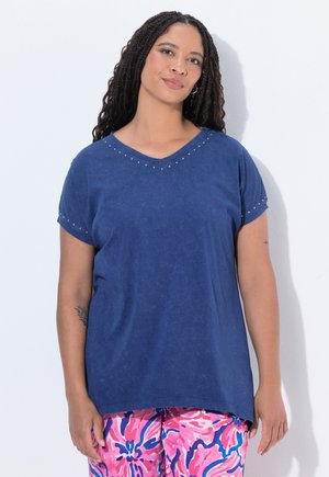 Femme aux longs cheveux tressés portant un t-shirt bleu clouté à col en V et un pantalon rose à motifs floraux, debout sur un fond blanc.