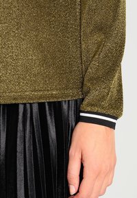 Nahaufnahme einer Hand neben einem goldglitzernden Langarmoberteil mit einem schwarz-weißen gestreiften Bündchen, kombiniert mit einem schwarzen plissierten Samröck.