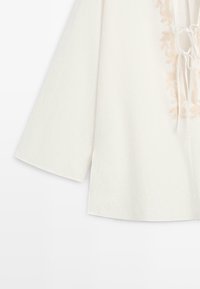 Chemise blanche à manches longues avec motif brodé beige et cordons près de l'encolure sur un fond uni.