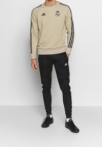 Uomo che indossa una felpa beige del Real Madrid con strisce nere Adidas, pantaloni della tuta neri Adidas, calzini bianchi e scarpe sportive nere.