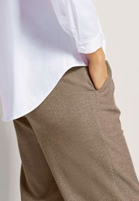 Witte langebloezende shirt met een afgeronde zoom, gecombineerd met getextureerde bruine broeken met zijzakken, weergegeven in een zijaanzicht.