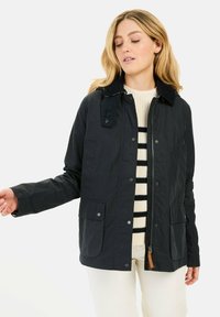 Sort, let jakke med en kordfløjlskrave, trykknapper, to forlommer og lynlås, båret over en stribet sweater.