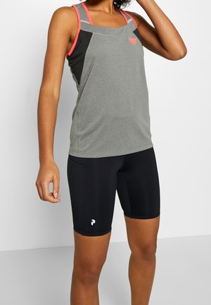 Kobieta ubrana w szary sportowy top bez rękawów z różowymi ramiączkami i czarne dopasowane sportowe spodenki, stojąca na białym tle.