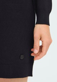 Vestido negro de punto que presenta un primer plano de una mano cerca de un botón negro en el dobladillo, mostrando una textura acanalada y mangas ajustadas.