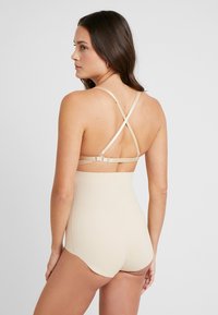 Hosszú, sötét hajú nő, bézs színű, magas derekú alakformáló alsóneműt és bézs színű melltartót visel, amelynek hátul keresztbe futó pántjai vannak.