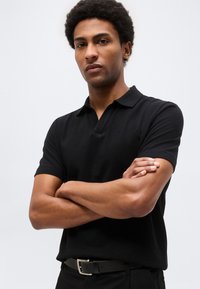 Νέος άντρας με σταυρωμένα χέρια, ντυμένος με μαύρο πόλο με κοντά μανίκια και μαύρη ζώνη, στέκεται μπροστά σε ένα απλό φωτεινό φόντο.