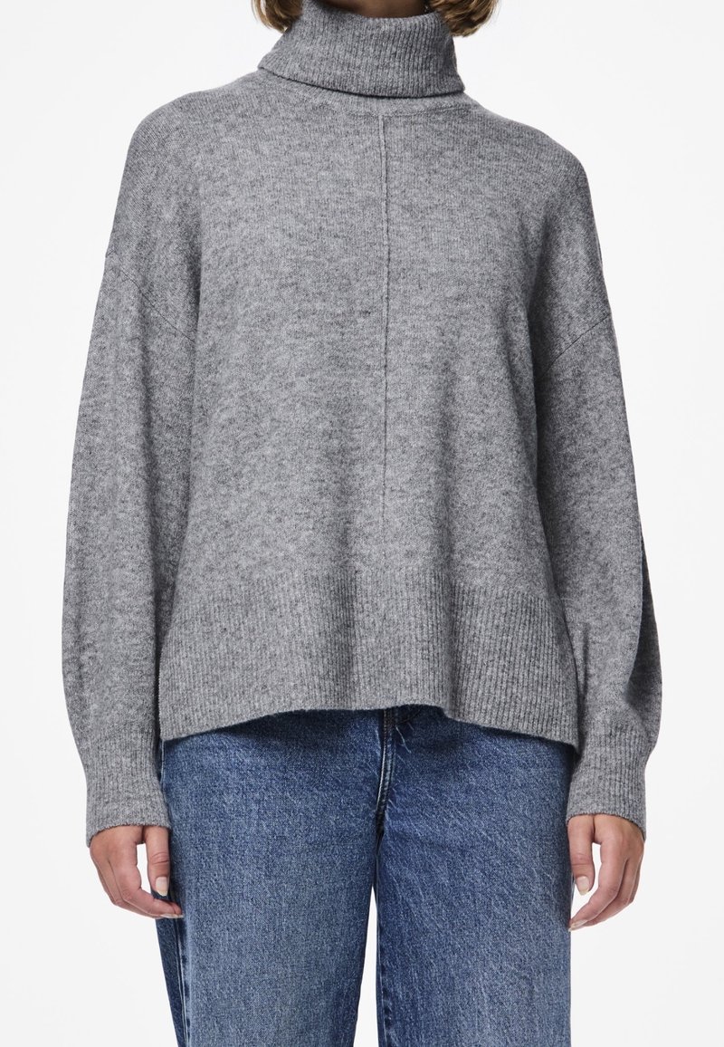 Femme portant un pull gris à col roulé ample avec un ourlet côtelé et un jean bleu taille haute, montrée du cou jusqu'aux hanches.