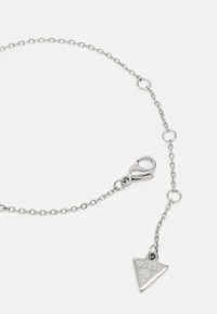 Silberne Metallkette Armband mit einem Karabinerhaken. Verfügt über einen angehängten dreieckigen Anhänger, der mit "GUESS?" und einem Fragezeichen graviert ist.