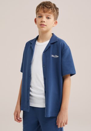 Chłopiec z krótkimi blond włosami, ubrany w niebieską krótko rękawową koszulę zapinaną na guziki na białą koszulkę i pasujące niebieskie spodnie, stojący na jednolitym tle.