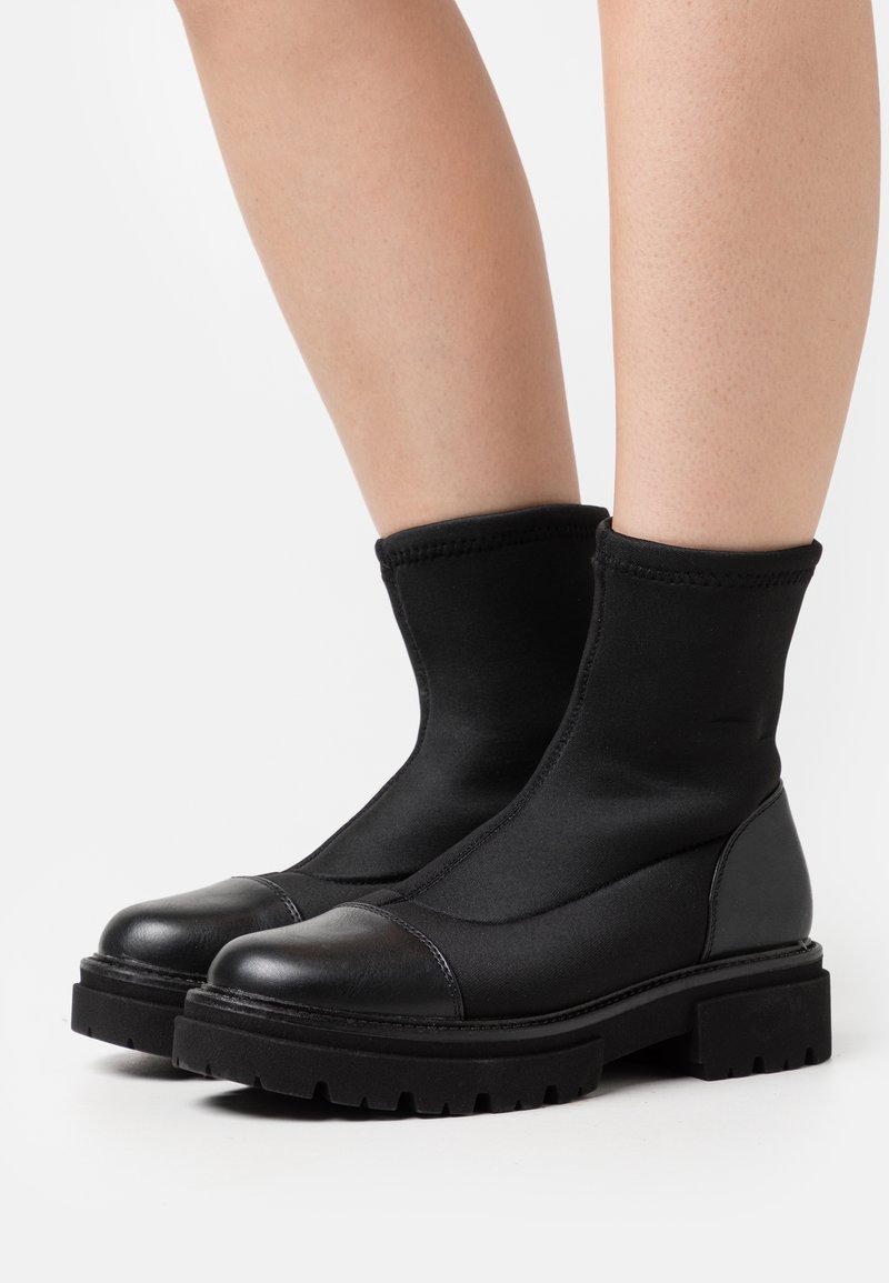 Botas negras de tobillo con suelas robustas y puntas de cuero liso, llevadas sobre piernas desnudas contra un fondo blanco.