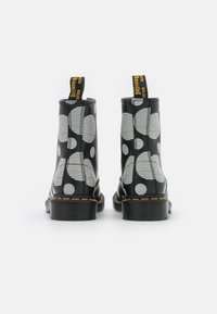 Stivaletti neri alla caviglia con finitura lucida, caratterizzati da motivi circolari bianchi e strisce orizzontali. Linguetta gialla e suola nera con cuciture visibili.