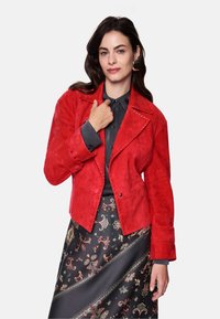 Red suede jacket with a notch collar, decorative stitching, and button closure. Worn over a dark blouse and patterned skirt.