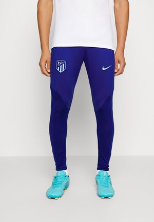 Homme portant un pantalon de sport bleu avec les logos Atlético Madrid et Nike, des chaussettes blanches et des chaussures de football bleu clair, debout sur un sol gris.