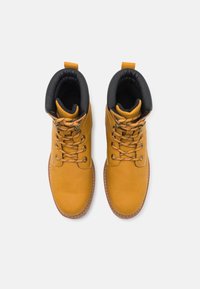 Botas de tobillo de cuero amarillo con cuello acolchado negro, ojales de metal y suela de goma marrón en contraste. Los cordones son de color canela con un ligero patrón.
