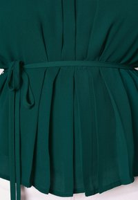 Top in poliestere teal con design arricciato e cintura regolabile, con pieghe sul retro e una texture liscia. L'orlo è leggermente curvo.