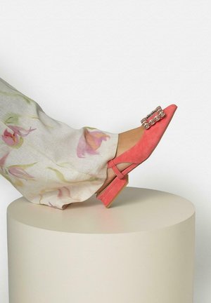 Been met bloemenbroek en koraalroze platte schoen met decoratieve gesp, rustend op een lichte ronde sokkel tegen een witte achtergrond.