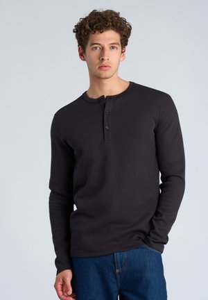 Junger Mann mit lockigem Haar, der ein schwarzes langärmeliges Henley-Shirt und blaue Jeans trägt, steht mit einer Hand in der Tasche vor schlichtem Hintergrund.