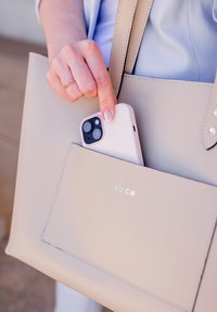 Béžová taška přes rameno s přední kapsou, která drží světle růžové pouzdro na telefon. Taška má hladkou texturu a stříbrné kovové detaily.