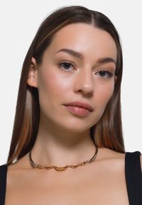 Metal choker halskæde med sammenflettede guld- og sølvbølge-designs, glat tekstur og en poleret finish.