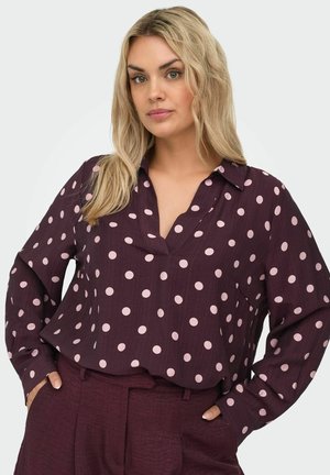 Blonde vrouw die een donkerpaarse blouse draagt met lichtroze stippen en bijpassende donkerpaarse broek, handen in de zakken, neutrale achtergrond.