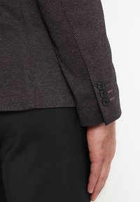 Dettaglio di un blazer grigio a spina di pesce con un polsino decorato da tre bottoni neri, tessuto testurizzato e una leggera accentuazione di colore visibile sulla manica.