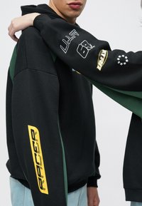 Kaks inimest, kellel on mustad ja tumerohelised dressipluusid kollase ja valge graafilise kirjaga varrukatel, ühevärvilise tausta ees.