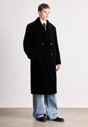 Zwarte, dubbelgeknoopte overjas gemaakt van textuurstof, met grote knopen en zijzakken, gedragen over lichte denim broek en zwarte schoenen.