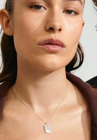 Collana con pendente d'argento su una catena fine, caratterizzata da un ciondolo rettangolare e testurizzato. Il modello indossa un indumento marrone scuro con un colletto sottile.