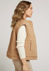 Gilet sans manches beige en tissu texturé, avec un col montant et deux poches avant, porté par-dessus un pull à manches longues blanc.