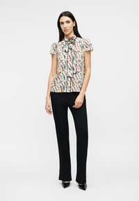 Kvinde iført en mønstret bluse med sløjfe og sorte bukser med lige ben, stående foran en ensfarvet baggrund.