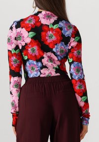 Vrouw draagt een langemouwen top met een rood, roze en blauwe bloemenpatroon op zwart, gecombineerd met hooggerokte donker bordeauxrode broek.
