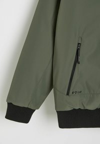 Oliwkowa kurtka o matowej, gładkiej fakturze, z zamkniętą kieszenią na zamek i czarnymi ściągaczami. Minimalistyczny design z detalem marki.