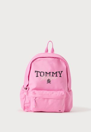 Rózsaszín Tommy Hilfiger hátizsák elülső cipzáras zsebbel és a márka fekete-fehér logójával világos egyszínű háttéren.