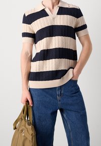 Pruhované pletené polo tričko v béžové a námořnické modři, s výstřihem do "V", krátkými rukávy a strukturovanou látkou. Kombinováno s modrými džínami a béžovou taškou.
