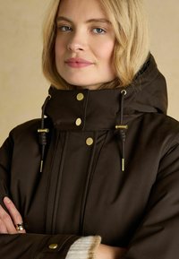 Blond naine siniste silmadega kannab tumedast pruunist kapuutsiga mantlit, millel on kuldsed nupud ja tõmblukud, risti käed.