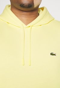 Felpa gialla in morbido tessuto, dotata di tasca a marsupio e cordini con terminali in plastica. Logo Lacoste ricamato sul petto.