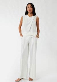 Biała bezrękawnik z drapowanym wzorem i ozdobnym guzikiem, połączony z luźnymi białymi spodniami i sandałami na otwartym nosku.