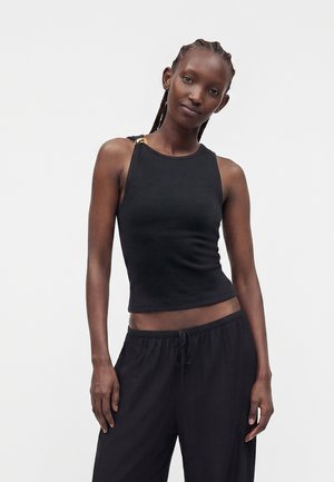 Jeune femme aux cheveux tressés porte un haut noir sans manches et un pantalon noir ample, debout devant un fond clair uni.