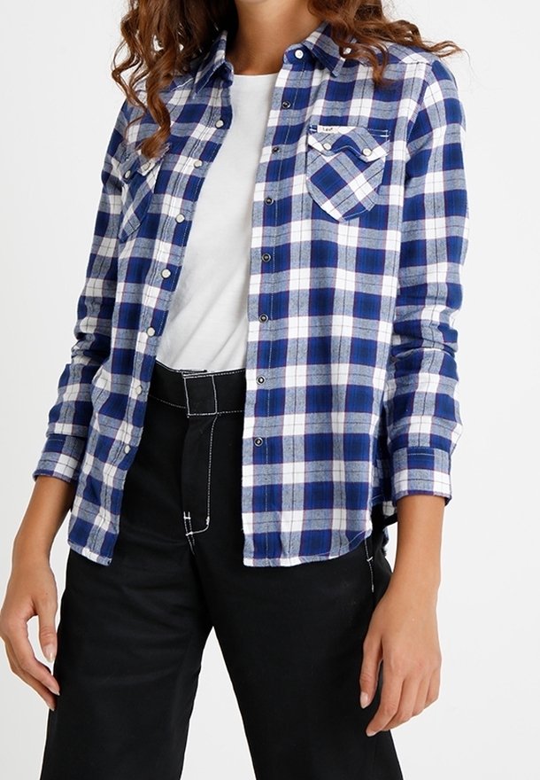 Personne portant une chemise à carreaux bleu et blanc boutonnée par-dessus un haut blanc, associée à un pantalon noir avec des coutures blanches visibles.