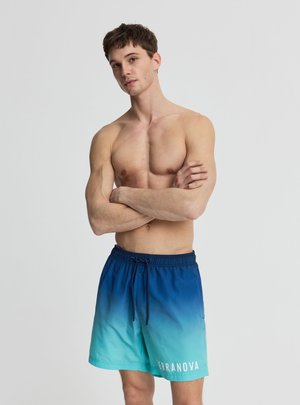 Giovane uomo senza maglietta con le braccia incrociate che indossa costumi da bagno sfumati dal blu al verde acqua con coulisse e scritta "TERRANOVA" sulla gamba sinistra.