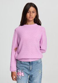 Maglione lavorato a maglia rosa chiaro con scollatura tonda e maniche lunghe, abbinato a jeans blu con testi grafici e accenti a forma di stella.