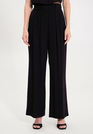 Femme portant un pantalon noir taille haute à jambes larges et des talons ouverts noirs, debout devant un fond clair uni.