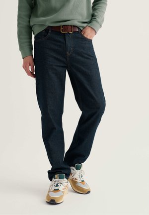 Person trägt dunkelblaue Jeans, einen grünen Pullover, einen braunen Gürtel und beige, weiße und grüne Sneakers, steht mit einer Hand in der Tasche.
